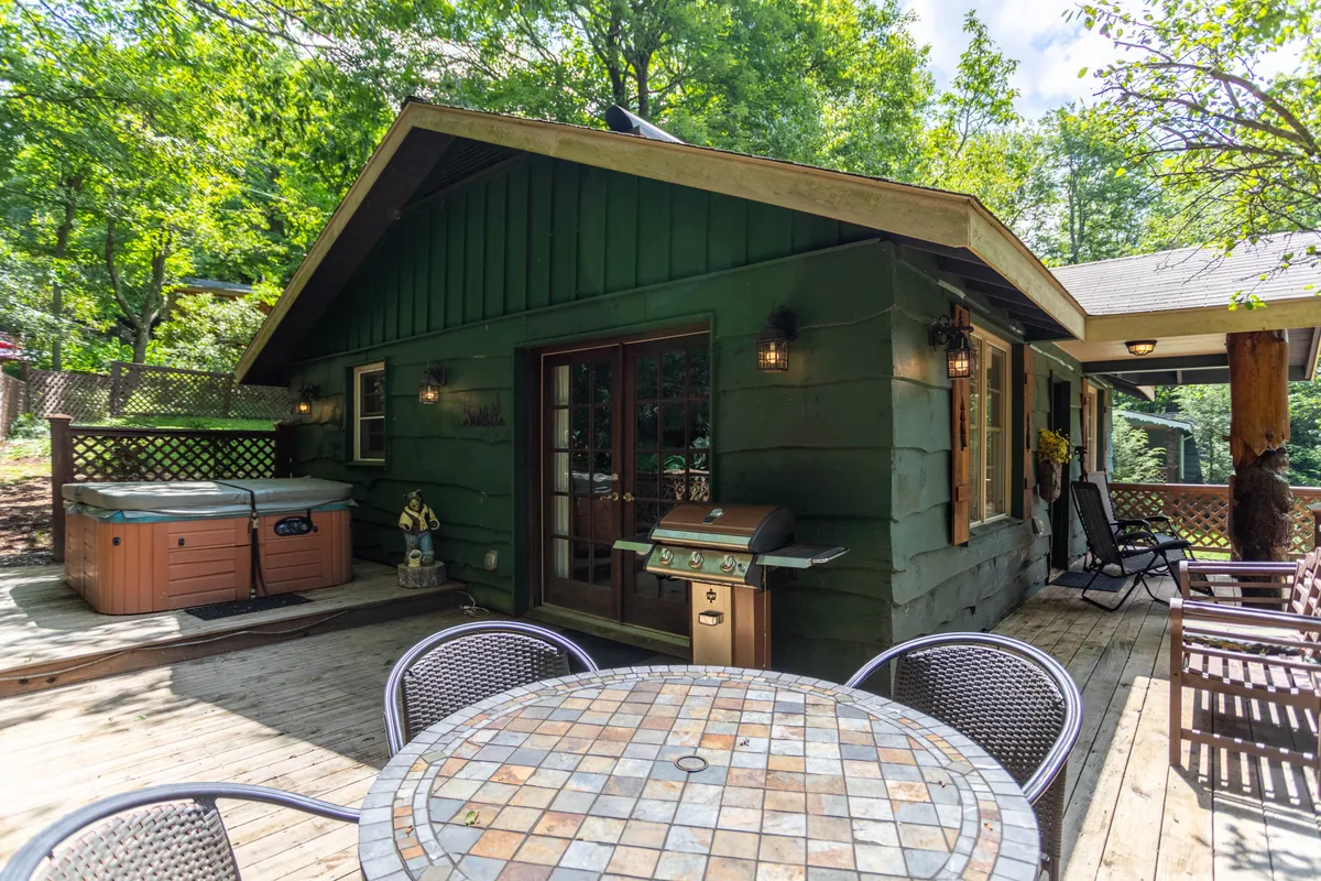This image presents a well-appointed wooden deck featuring a Hot Springs hot tub, outdoor dining table, and a gas grill. The setting is surrounded by lush greenery, creating a private and inviting outdoor living area ideal for relaxation, dining, and socializing.