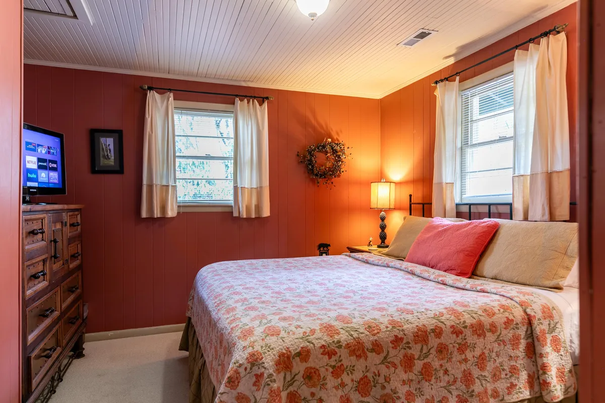 A cozy bedroom featuring a bed with floral bedding, a wooden dresser, a window with curtains that lets in natural light, and a table lamp. The inviting ambiance is accentuated by the soft glow of the lamp and the comfortable, high-quality linens.