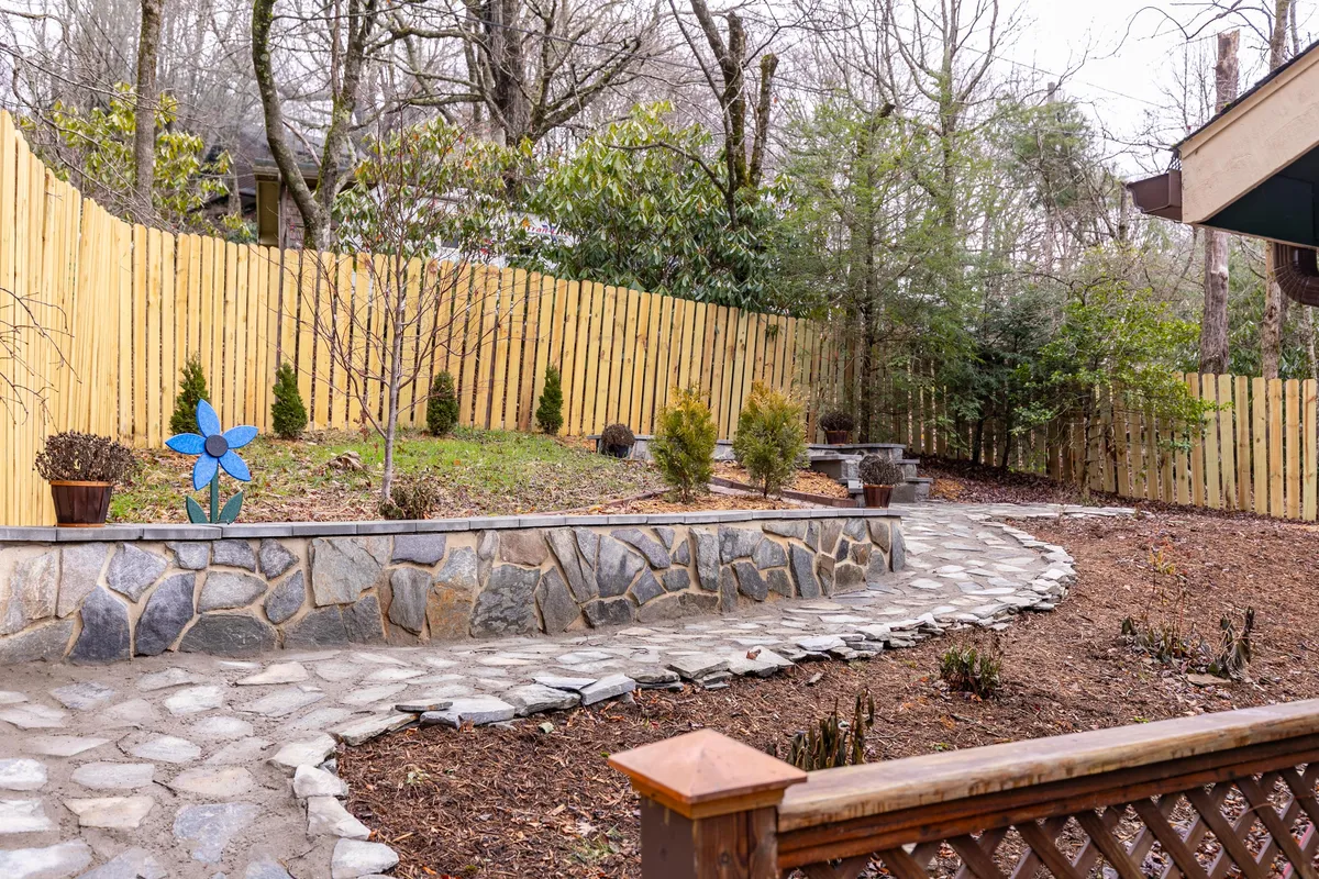 This exterior image showcases a serene stone pathway bordered by lush hedges, a whimsical blue flower sculpture, a sturdy wooden fence, and a welcoming outdoor sitting area. The fenced area is prominent, suggesting a safe and pleasant space for pets to roam while owners relax and enjoy the scenery.