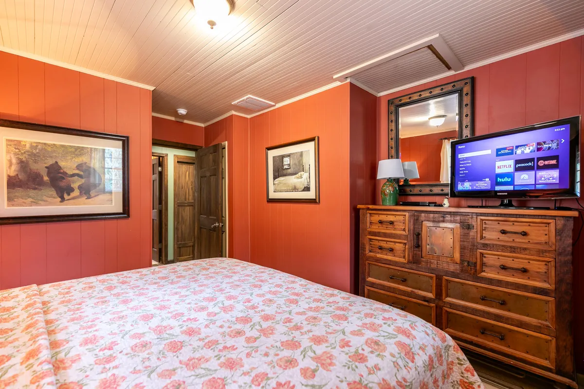 A warmly decorated bedroom with a floral patterned bedspread, wooden dresser, framed artworks above the bed, and a television set up for in-room entertainment. The soft lighting and comfortable atmosphere highlight the room's relaxing features.