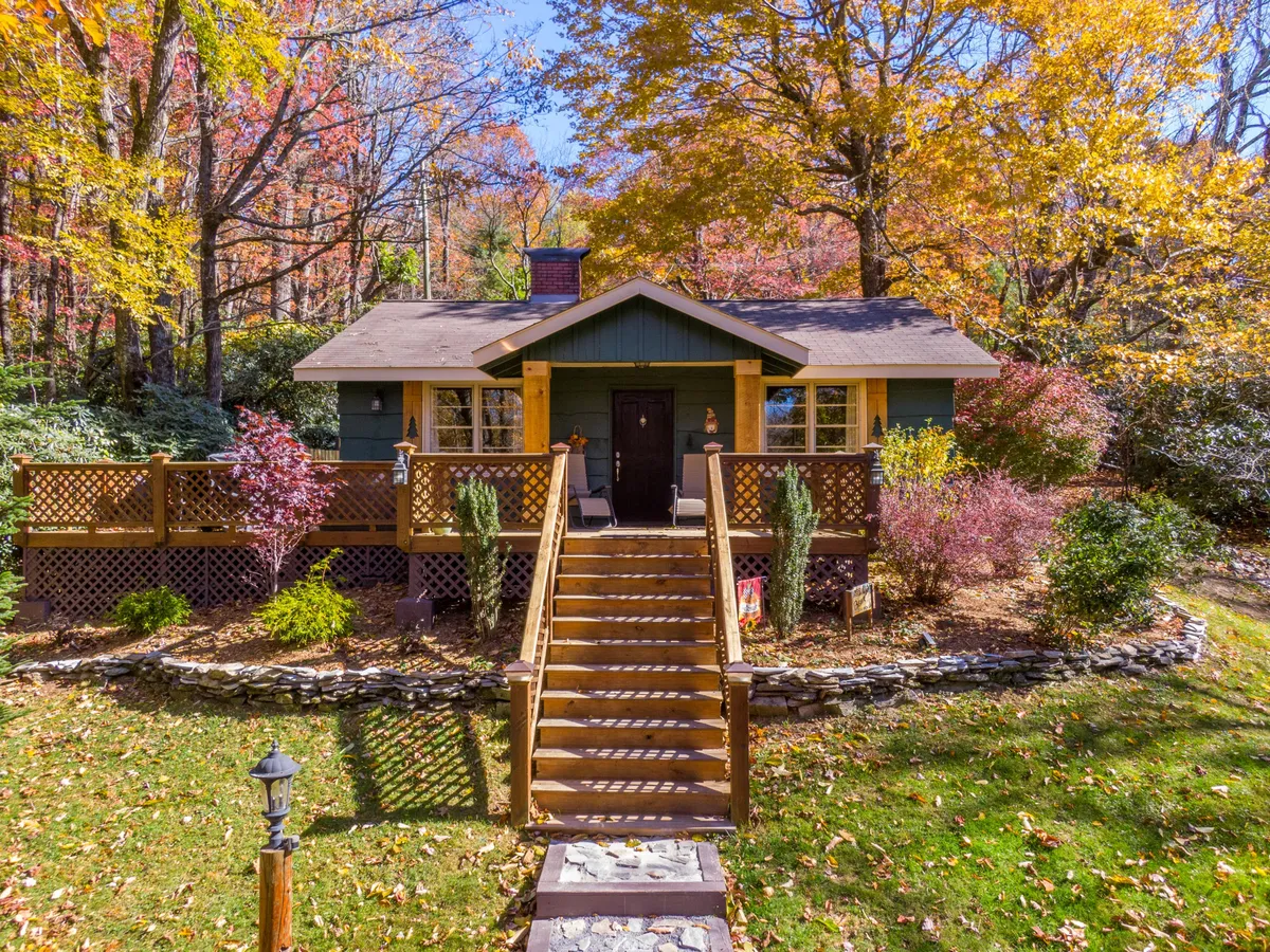 The image showcases the welcoming front porch of a cozy cabin nestled amid vibrant, colorful foliage and flowering plants. The staircase leads to an inviting porch, perfect for relaxing while surrounded by the lush rhododendron grove. The scene radiates warmth, privacy, and the beauty of nature—ideal for enjoying morning coffee or sunset views right outside the cabin door.