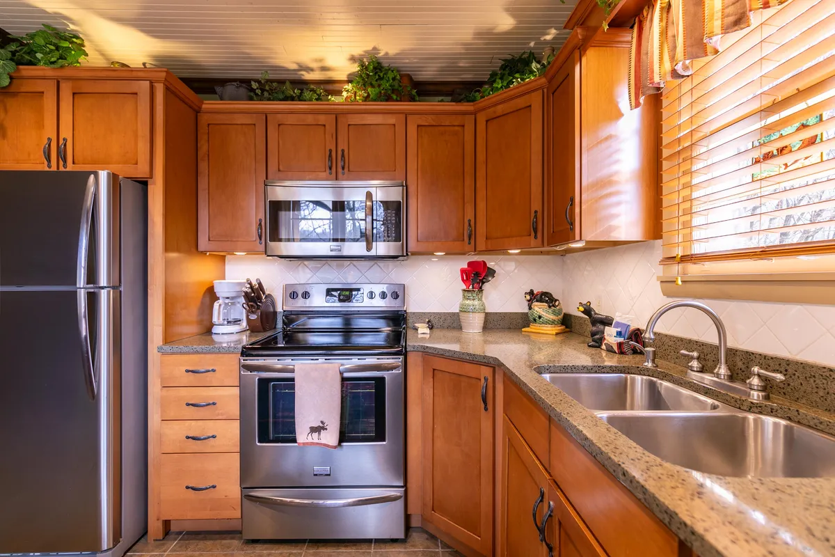 This image features a cozy kitchen scene with stainless steel appliances, granite countertops, rich wooden cabinets, and a kitchen sink beneath a window with blinds. The warm wood tones, sleek surfaces, and natural light offer a comprehensive view of the kitchen’s upscale finishes, matching the description of solid maple cabinetry and granite surfaces. The presence of various appliances and the sink showcases the kitchen's functionality.