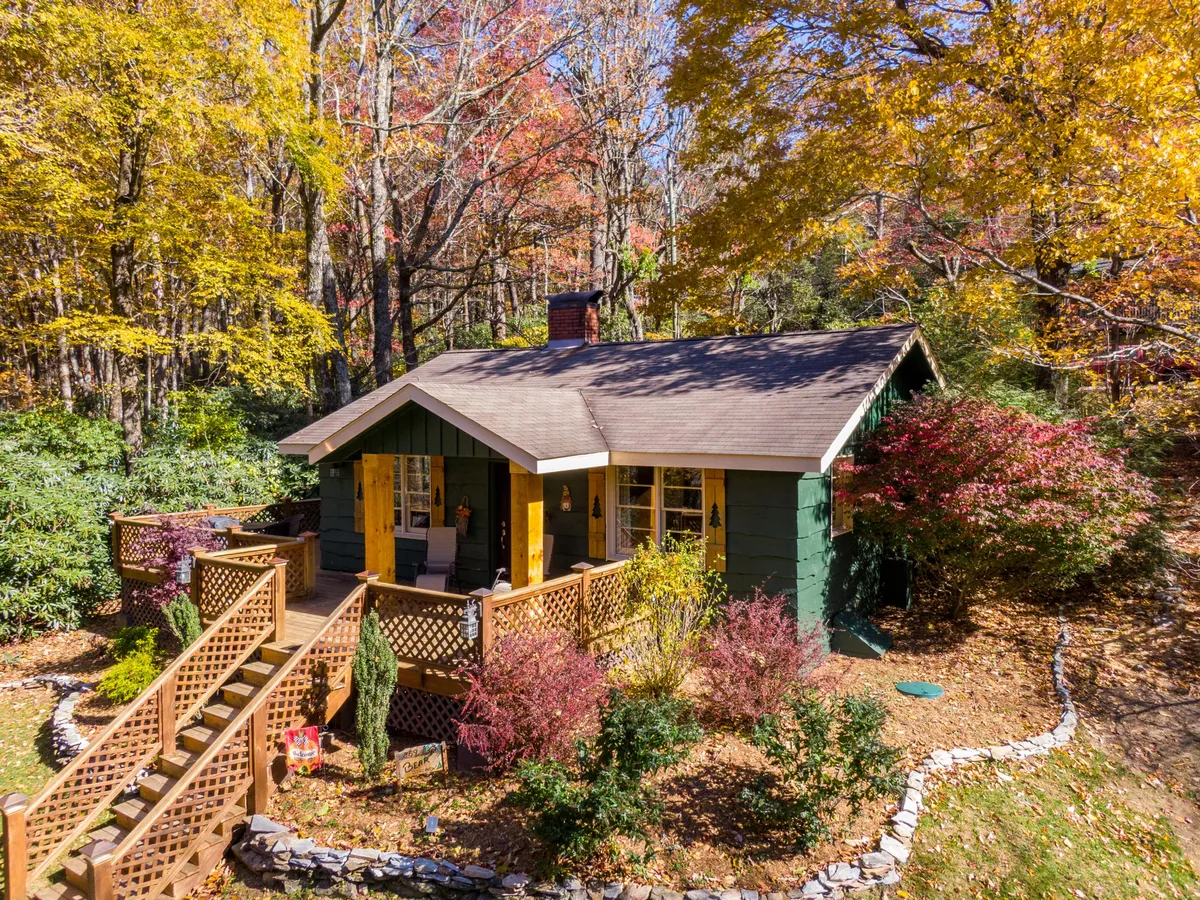A charming green cottage nestled among vibrant autumn foliage, with a welcoming wooden porch and comfortable seating. The surrounding garden and colorful shrubs add to the inviting, cozy ambiance.