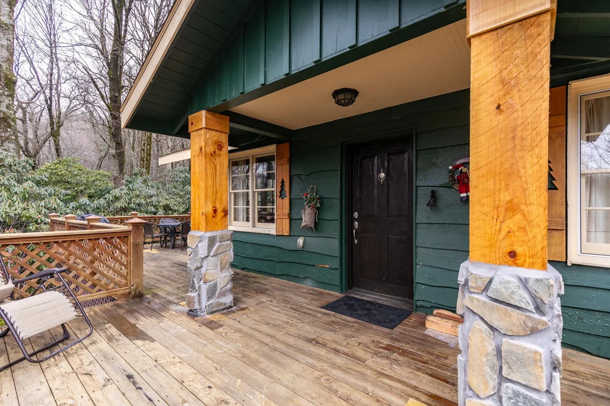 The image showcases the front door of the cottage with wooden pillars, stone accents, and a spacious deck area. The scene is welcoming and hints at the seamless indoor-outdoor lifestyle.