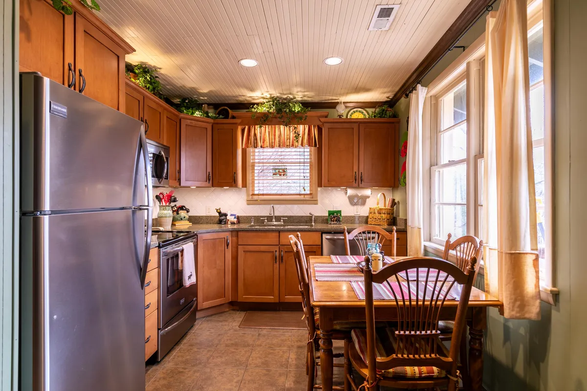 A broad view of the kitchen and dining area, featuring a central kitchen island, a dining table set for meals, large windows letting in natural light, and visible appliances. The open design connects the kitchen and dining spaces, echoing the narrative about togetherness and seamless flow between cooking and socializing. Accent lighting helps create a warm, welcoming mood.