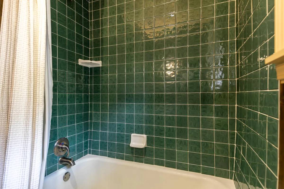 A tranquil bathroom with green tiled walls, a white shower curtain, and a spacious bathtub. The atmosphere is peaceful, perfect for soothing sore muscles after an active day.